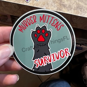 May include: A round sticker with a black border and a green background. The sticker features a black cat paw with red claws and a red paw pad. The text "Murder Mittens Survivor" is written in red around the paw.