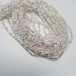 May include: A close-up of a silver beaded necklace. The beads are small and square-shaped, and they are strung together in a long strand. The necklace is coiled up on a white surface.