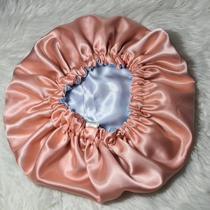 Puede incluir: Una gorro de dormir de satén de color melocotón con un forro de satén azul claro. El gorro está fruncido en la parte superior y tiene un cordón.