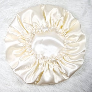 May include: Ivory satin bonnet with a gathered elastic edge. The bonnet is made of a smooth, shiny fabric and is designed to protect hair while sleeping. The bonnet is a light cream color and has a circular shape.