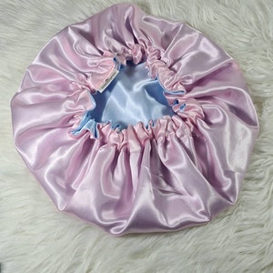 May include: A pink satin bonnet with a light blue satin lining. The bonnet is gathered at the top and has a small label with the text "Satin Bonnet" on it.