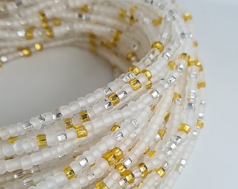 May include: A white beaded waist bead with gold and silver accents. The beads are arranged in a circular pattern and are strung on a clear thread.