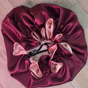 May include: A reversible satin hair bonnet featuring a deep burgundy exterior and a soft champagne-colored interior. The circular accessory has a gathered, pleated design with a black adjustable drawstring cord and a small white brand tag.