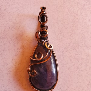 Könnte beinhalten: Ein tropfenförmiger Anhänger mit einem dunkelvioletten Edelstein, der in Kupferdraht gewickelt ist. Der Draht ist verdreht und zu dekorativen Wirbeln und Schleifen geformt.