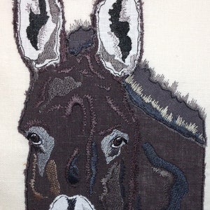 May include: Embroidered donkey head with brown, gray, and white fabric. The donkey's eyes are stitched with blue thread.