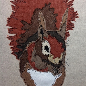 May include: A brown and red squirrel with a bushy tail, embroidered on a beige fabric background.