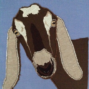 May include: A brown and white goat with large, floppy ears. The goat is looking directly at the camera with a curious expression. The goat is made of fabric and has a stitched-on appearance.