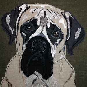 Puede incluir: Primer plano de una imagen bordada de la cara de un perro. El perro tiene un pelaje marrón claro y blanco con marcas negras alrededor de los ojos y la nariz. Los ojos del perro son marrones y la nariz es negra.
