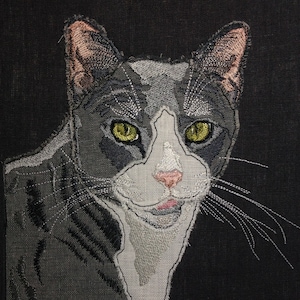 Könnte beinhalten: Gesticktes Katzenporträt auf schwarzem Stoff. Die Katze hat grau-weißes Fell, gelbe Augen und eine rosa Nase. Die Stickerei zeigt die Merkmale der Katze, einschließlich Schnurrhaare und Ohrendetails.