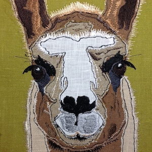 Könnte beinhalten: Eine Nahaufnahme eines braunen und weißen Lamas, das auf einen grünen Stoffhintergrund gestickt ist. Das Gesicht des Lamas ist detailliert gestickt, einschließlich seiner Augen, Nase und seines Mundes.