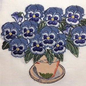 Può includere: Un bouquet di violette ricamate in blu e bianco in un vaso marrone con un sottopiatto bianco.