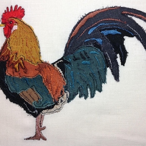 May include: A colourful rooster embroidered on a white fabric background. The rooster is made of different fabric scraps sewn together, creating a patchwork effect. The rooster's tail feathers are long and flowing, and its comb is red.