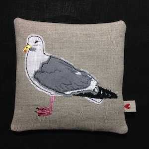 May include: A gray and white embroidered seagull on a beige linen pillow with a red heart tag.