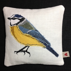 May include: A white linen pillow with an embroidered bluebird with yellow and blue accents. The bird is facing right and has a small red heart on the bottom right corner.