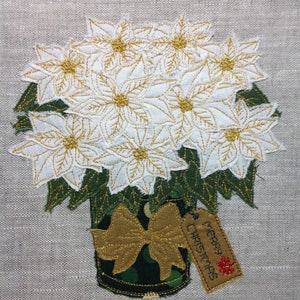 Può includere: Fiori di stella di Natale bianchi con dettagli di glitter dorati in un vaso verde con un fiocco dorato e un'etichetta che dice "Buon Natale".