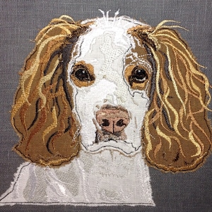 May include: Embroidered image of a white and brown spaniel dog with long, flowing ears. The dog is looking directly at the viewer with a gentle expression.