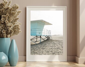 Könnte beinhalten: Ein weiß gerahmtes Foto eines hellblauen Rettungsschwimmers auf einem Sandstrand. Der Stand hat eine Holzleiter und ein Metallgeländer. Der Strand ist leer und der Himmel ist klar.
