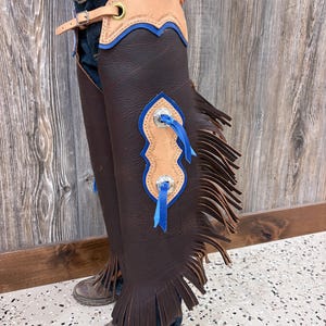 May include: Brown leather chaps with blue and tan accents. The chaps feature fringe along the bottom and decorative blue ribbon. The top has a tan leather section with blue trim. A silver buckle and belt are visible.