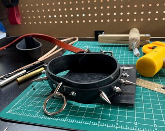 May include: Black leather collar with silver spikes and a large silver ring. The collar is on a green cutting mat with a ruler and yellow tool in the background. A leather strap is also visible.