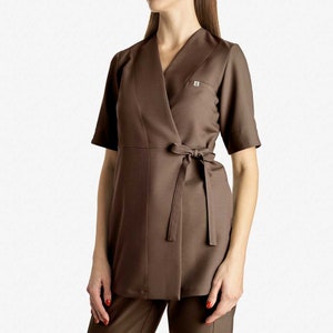 May include: A brown wrap-style tunic with a V-neckline and a tie belt. The tunic has short sleeves and a small logo on the left chest.