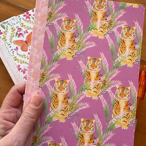 May include: A notebook with a purple cover featuring a repeating pattern of orange tigers and green foliage. A second notebook with a floral and butterfly design is partially visible. An orange pen is in the background.