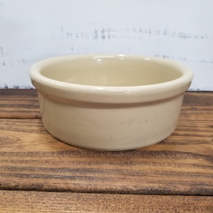 May include: A round, cream-colored ceramic bowl with a smooth, glossy finish and a thick, rounded rim. The bowl features a simple, open design with a deep interior and a flat base, showcasing a uniform, neutral tone throughout its entire structure.
