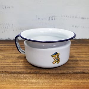 May include: A white enamel chamber pot with a dark blue rim and a matching handle. The exterior features a small, colorful graphic of a brown teddy bear wearing a blue hat and a yellow shirt, depicted in a walking pose on the side of the vessel.