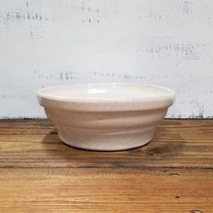 May include: A round, off-white ceramic bowl with a speckled texture and a wide, flat rim. The bowl features a series of horizontal ridges along its exterior side, creating a tiered profile that tapers slightly toward the base.