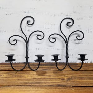 May include: Two black metal wall sconces with a decorative, twisted design. Each sconce has three candle holders and a swirling, vine-like top. The sconces are displayed on a wooden surface against a white background.