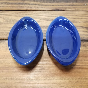 May include: Two cobalt blue, oval-shaped ceramic dishes. The dishes have a smooth, glossy finish and are set against a wooden background. The design is simple and elegant, suitable for serving appetizers or small portions.