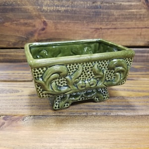 May include: A green ceramic planter with a rectangular shape and ornate floral detailing. The planter has a textured surface and sits on small feet. The interior is a darker shade of green.