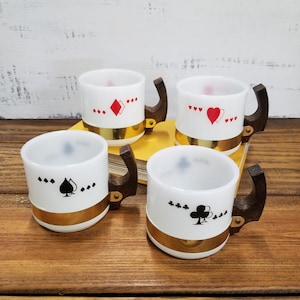 May include: A set of four white glass mugs featuring playing card suit symbols. Each mug has a dark wooden handle, a gold band around the base, and a specific suit icon: a red diamond, a red heart, a black spade, and a black club.