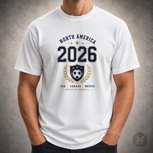 May include: A white short-sleeved t-shirt featuring a graphic with the text NORTH AMERICA 2026, USA CANADA MEXICO, and UNITED BY FOOTBALL. The design includes a central shield with a soccer ball, gold laurel branches, and three gold stars.