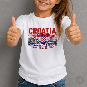 Może przedstawiać: Biały t-shirt z kr&oacute;tkim rękawem z nadrukiem graficznym zawierającym napis CROATIA, flagę Chorwacji w kształcie serca, ilustrację miasta nadmorskiego, most oraz napis Ponos U Srcu 20 26 z małą ikoną piłki nożnej umieszczoną pod sercem.