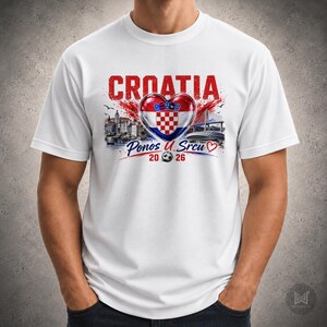 Może przedstawiać: Biały t-shirt z kr&oacute;tkim rękawem z nadrukiem graficznym zawierającym czerwony napis CROATIA, chorwacką flagę w kształcie serca, ilustrację miasta nadmorskiego, most oraz napis Ponos U Srcu 2026 z małą ikoną piłki nożnej.