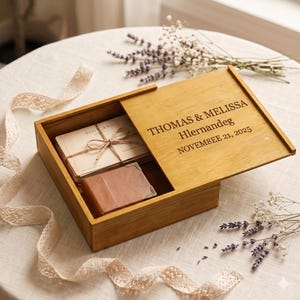 May include: A wooden box with a sliding lid, engraved with names and a date. Inside, postcards tied with twine and a small leather wallet are visible. The box is open, revealing its contents, and sits on a white surface with lace trim and floral accents.