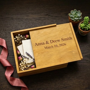 May include: A wooden box with a sliding lid, engraved with the names "Anna & Drew Smith" and the date "March 10, 2026." The box is partially open, revealing white cards, dried flowers, and lavender. A pink ribbon and small potted succulents are also visible.