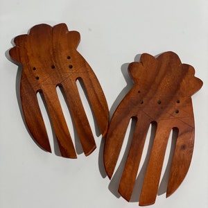 May include: Two wooden salad servers. Each server has a handle with a scalloped edge and four tines. The wood is a warm brown color, and the servers are designed for tossing and serving salads.