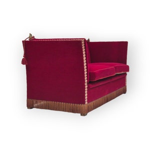 Puede incluir: Un sofá de terciopelo rojo con ribete de flecos y detalles dorados. El sofá tiene un respaldo capitoné y un color rojo intenso.