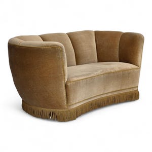 Könnte beinhalten: Ein geschwungenes Zweisitzer-Sofa, bezogen mit einem weichen, beigefarbenen Stoff. Das Sofa hat eine abgerundete Rückenlehne und Armlehnen sowie einen dekorativen Fransenbesatz am Sockel. Das Design erinnert an Vintage-Möbel.