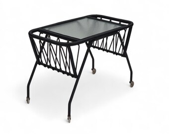 May include: Black metal side table with a glass top and wheels. The table has a woven design on the sides.