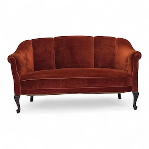 Puede incluir: Un sofá de tres plazas de estilo vintage en terciopelo color óxido. El sofá presenta un respaldo curvo, brazos enrollados y patas de madera oscura. El diseño es clásico y elegante, adecuado para una sala de estar. Mide unos 200 cm de largo.