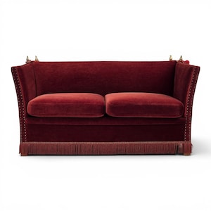 May include: A deep red velvet sofa featuring two seat cushions, high flared armrests, and a base adorned with a matching fringe trim. The front edges of the armrests and the lower frame are detailed with a row of small metallic nailhead studs.
