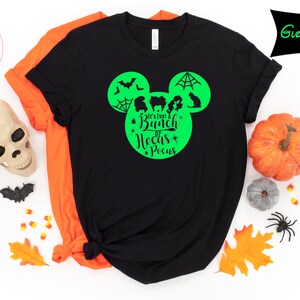 May include: Black t-shirt with a green graphic design featuring a Mickey Mouse head shape with bats, a spiderweb, and silhouettes of characters. The text "It's Just a Bunch of Hocus Pocus" is in the center. An orange shirt is visible behind it. Halloween theme.