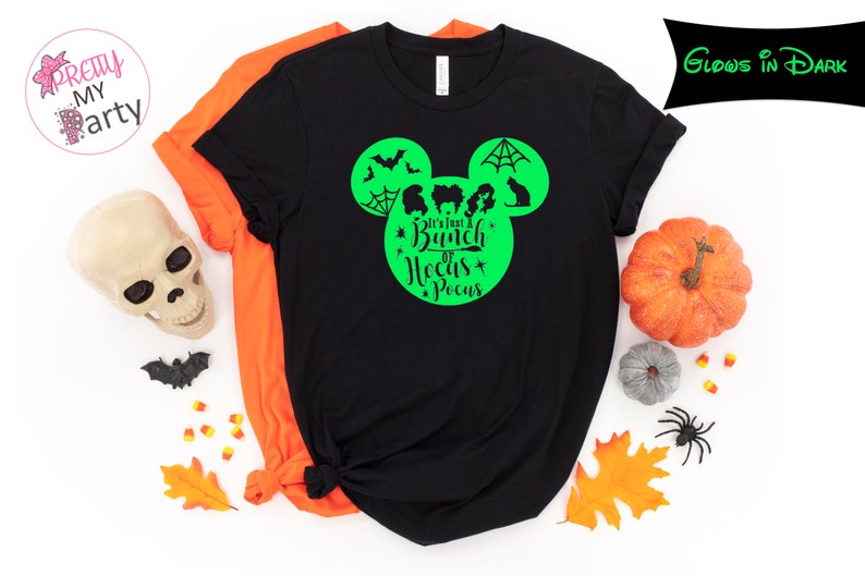 May include: Black t-shirt with a green graphic design featuring a Mickey Mouse head shape with bats, a spiderweb, and silhouettes of characters. The text "It's Just a Bunch of Hocus Pocus" is in the center. An orange shirt is visible behind it. Halloween theme.