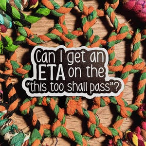 May include: A white sticker with black text that reads "Can I get an ETA on the 'this too shall pass'?" is centered on a colorful woven background of orange, green, and other colors.