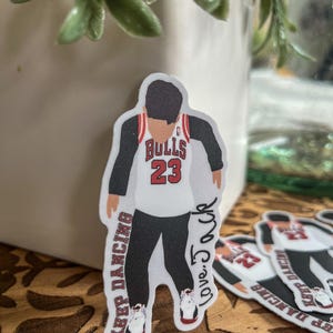 May include: A sticker of a basketball player wearing a white jersey with the words "Bulls 23" on the front. The player is wearing black pants and red and white sneakers. The sticker has the words "Keep Dancing" and "Love. J.O.C.R" written on it.