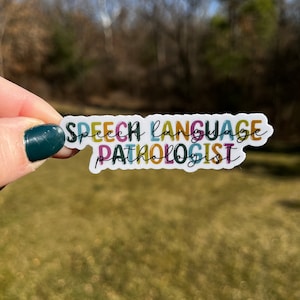 May include: A colourful sticker with the words "Speech Language Pathologist" in various fonts and colours. The sticker is held up against a blurred background of trees and grass.