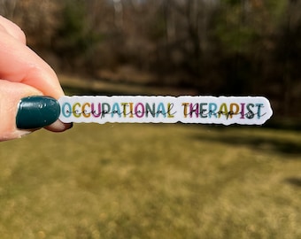 Può includere: Un adesivo bianco con il testo "Occupational Therapist" in lettere colorate. L'adesivo è tenuto in una mano con smalto per unghie verde scuro su uno sfondo sfocato di alberi ed erba.