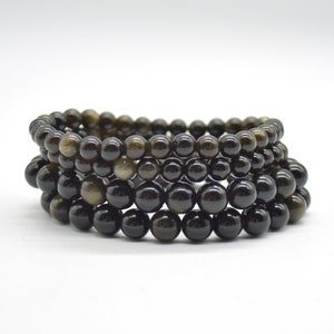 May include: A stack of four black beaded bracelets with a gold shimmer. The bracelets are made of round beads and are stacked on top of each other.